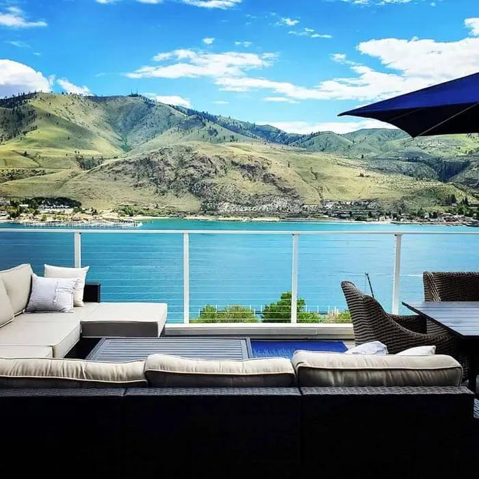 Backyard outdoor living space with gray composite decking and white horizontal cable railing overlooking a blue lake and mountains in the background.